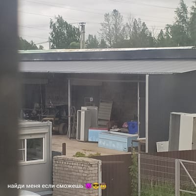The image shows an outdoor scene with a large, dark-colored building that appears to be a workshop or storage facility. The building has a corrugated metal roof and a wide open entrance. Inside the building, various objects are visible, including what looks like machinery, shelving units, and what could be appliances or large containers. There is also a small, light-colored structure with a window to the left of the main building. A brick wall and a metal fence are in the foreground. The sky is overcast. Text in the lower left corner of the image reads "найди меня если сможешь" which translates to "find me if you can", followed by three emojis: a devil face, a sunglasses-wearing face, and a cowboy face. The overall impression is of an industrial or utilitarian area, possibly a junkyard or a place where items are stored or repaired. This scene is not specifically identifiable as being in Pskov, Russia, based solely on the visual information provided.