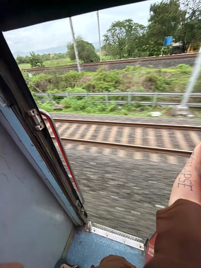 The image shows a view from a moving train, likely in the vicinity of Saphale, India, given the context. The perspective is from inside the train, looking out a doorway. The tracks of the train are visible in the foreground, blurred due to motion. Beyond the tracks, a lush green landscape with trees and distant hills can be seen under an overcast sky. A person's arm with a tattoo is in the lower right corner of the frame, indicating their presence in the scene. The interior of the train, including a door frame and a metal handle, is visible on the left side. The overall impression is of a journey through a rural or semi-rural area.