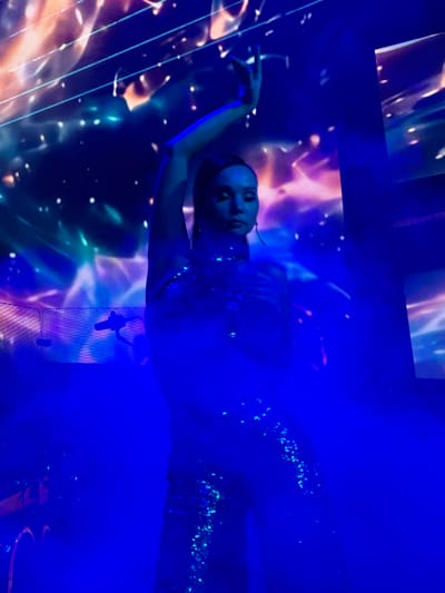 The image depicts a female performer on a stage, bathed in blue and purple light, with a vibrant, abstract light show serving as the backdrop. Her face is illuminated, and her arm is raised, suggesting a dance or performance. She is wearing a shimmering, sequined outfit that catches the light. The stage appears to be set up for a concert or similar event, with some equipment visible in the background. The overall atmosphere is dynamic and theatrical. The location in Akhalubani, Georgia is not discernible from the image itself.