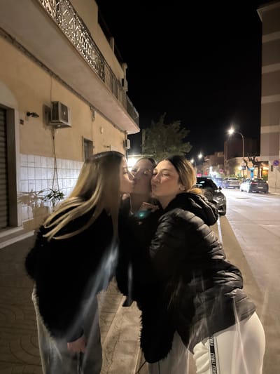 The scene depicts three young adult females at nighttime on a paved sidewalk in Stornara, Italy. They are arranged in a line from left to right, engaged in a chain of kisses. The leftmost individual, with long blonde hair and wearing a black textured jacket over light-colored pants, is kissing the middle individual on the cheek/mouth. The middle individual, with dark hair and wearing a dark top, is simultaneously being kissed by the leftmost person and kissing the rightmost individual on the cheek/mouth. The rightmost individual, with brown hair featuring blonde highlights pulled back, is wearing a black puffer jacket and white sweatpants with a repeated "miu miu" logo on the outer seam of the left leg, and is being kissed by the middle individual.

The immediate background on the left features a light-colored stucco building with a decorative wrought-iron balcony railing on an upper floor. Two external air conditioning units are mounted on the wall, below which a white tiled section is visible. A building number "32" is affixed to the wall. Shadows from an unseen object are cast onto the tiled wall. To the right of the subjects, a street runs parallel to the sidewalk, illuminated by active streetlights. Several vehicles, some parked and others possibly moving, are visible in the distance with their lights on. A tree with sparse foliage stands further back from the street. The sky is dark, indicating night.