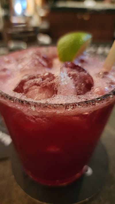 The image displays a close-up view of a deep red or magenta beverage in a glass, located in Jackson, United States of America. The drink contains multiple ice cubes and presents a frothy, bubbly surface. The rim of the glass is coated with a white, granular substance, consistent with salt or sugar. A green lime wedge serves as a garnish, positioned on the rim or floating on the surface of the liquid. A light-colored straw is partially visible on the right side, submerged in the drink. The glass rests on a dark, circular coaster. The background is significantly blurred, showing diffuse light sources and indistinct shapes, indicative of an indoor, commercial setting such as a bar or restaurant. No individuals, specific actions, or direct interactions are discernible within the frame.