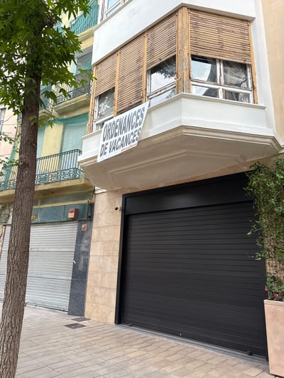 The image shows a street scene in the city of Igualada, Spain. In the foreground, a large black roller shutter door is visible, covering the ground floor of a building. To the left of the roller shutter is a light-colored tiled wall, and to the left of that, a silver roller shutter door. A mature tree with green leaves stands on the sidewalk to the left. Above the black roller shutter, a white balcony with weathered wooden shutters is visible. A banner with text hangs from the balcony, reading "ORDENANCES DE VACANCES" and "#ProuSor". Above this balcony, another building with a green-painted balcony and yellow and light blue painted walls is partially visible. The environment appears to be urban, with paved sidewalks and buildings lining the street.