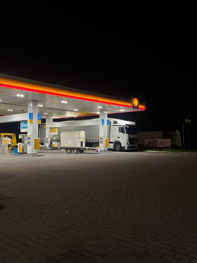 At night, at a Shell gas station in Horbów-Kolonia, Poland, a white semi-truck is parked at a diesel pump under the illuminated canopy. The truck is facing towards the right of the frame, and its trailer is partially visible. In front of the truck, on a trailer, is a large white rectangular container, possibly for AdBlue. A yellow tow truck is parked to the left of the diesel pumps, with a person in a white t-shirt and shorts standing near it, facing away from the camera. The gas station canopy is lit by overhead lights and features red and yellow accents. The ground is paved with interlocking concrete blocks. A billboard with an advertisement is visible in the background, as well as a green flag. The sky is dark and clear.