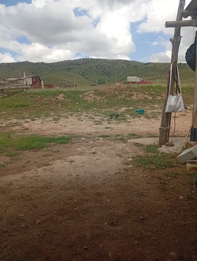 The image depicts a rural, mountainous landscape in Көксаяқ ауылдық округі, Kazakhstan, under a partly cloudy sky. In the foreground, a dirt path leads into a yard with scattered rocks and sparse green vegetation. To the left, a wooden fence borders a grassy area with a small, red-roofed building or shed. In the middle ground, a truck and a red car are parked on a dirt road. The background features rolling green hills dotted with trees, leading up to a rugged mountain range. On the right side of the image, a wooden post is secured with rope and has a white bag and a black bag hanging from it. A shovel and a bucket are also visible in the yard. The scene conveys a sense of rustic, pastoral life in a remote, natural setting.