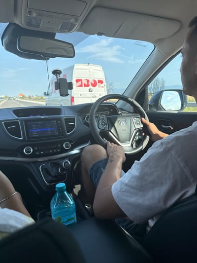 A first-person perspective shot from inside a car driving on a highway. The driver, a male with tattoos on his left forearm, is visible from the shoulders down, wearing a white t-shirt and denim shorts. His hands are on the steering wheel. In the foreground, a bottle of water is placed in the center console. The dashboard displays the radio interface, with the time at 17:22. Outside the car, a white van with red lettering is visible in front, along with other vehicles and a clear sky. The scene appears to be set in Shtarkovo, Bulgaria, based on the provided location information.