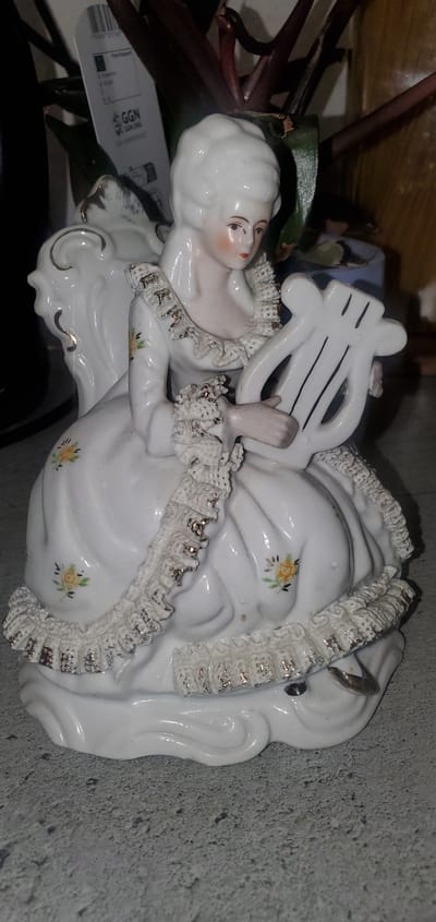 The image shows a close-up of a porcelain figurine of a woman in historical dress, holding a lyre. The figurine is white with delicate floral patterns and gold accents. It is placed on a gray, textured surface, possibly a countertop or table. In the background, there is a blurred plant with dark leaves and a price tag with German text, including "GGN.ORG". The figurine's details, such as her hair, face, and the ruffles on her dress, are clearly visible. The lighting is somewhat dim, casting soft shadows. This image was taken in Sasbach, Germany.