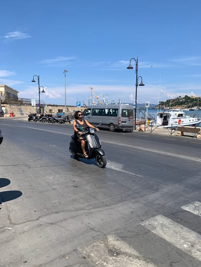 A person is riding a dark-colored scooter on a paved road. They are wearing a black tank top, dark shorts, sunglasses, and sandals. The scooter is a modern design with a headlight and handlebars. The scene is outdoors in a harbor area, with boats docked in the background and a crane visible further down. A grey van is parked behind the scooter, and a row of other parked scooters is visible to the left. The road has a pedestrian crossing painted on it. The sky is bright blue with some light clouds, suggesting a sunny day. The overall mood appears cheerful and relaxed, with the rider smiling and seemingly enjoying the ride. The time of day appears to be daytime, likely late morning or early afternoon. Location cues such as the boats, harbor setting, and the architecture of the buildings in the distance, along with the context provided, point to Porto Santo Stefano, Italy. No legible text is visible on the person or the scooter, but license plates are present on the van.