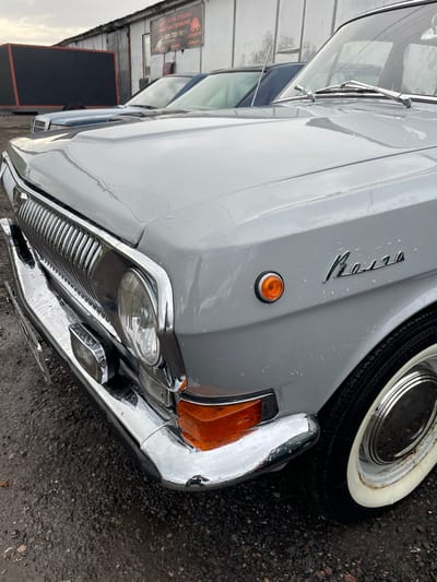 The image shows the front of a grey Volga car. The car has a chrome bumper and grill. There is a logo on the front fender that reads "Volga" in Cyrillic script. The car appears to be wet, likely due to rain. The location is unknown; the city of Khar'ino, Russia, is not directly indicated in the image itself.