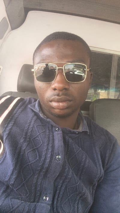 A man is depicted in a close-up, chest-up shot, likely a selfie, taken inside what appears to be a vehicle. He has dark skin, short, dark hair, and subtle facial hair, including a mustache and a goatee. His eyes are obscured by a pair of gold-framed sunglasses with dark, reflective lenses. The reflections in the sunglasses vividly capture an outdoor street scene, featuring several vehicles, including a prominent white car, and some trees or foliage, indicating that the man is either a driver or a passenger in a moving or parked vehicle.
He is wearing a dark blue, long-sleeved shirt or cardigan. The garment has a collared neckline and a distinct textured pattern across its surface, resembling a cable knit or an embossed design with diamond shapes and vertical ridges. Two dark, round buttons are visible on the front placket of his shirt. A black and white striped strap, which appears to be a seatbelt, diagonally crosses his left shoulder and chest area.
The setting behind him further confirms his presence in a vehicle. A dark headrest is visible directly behind his head, and the light-colored ceiling or roof interior of the vehicle is seen above him. To the right side of the frame (viewer's left), a dark panel or part of the vehicle's interior is visible. To the left side of the frame (viewer's right), a window offers a blurred view of the outside environment, consistent with being inside a vehicle. The overall lighting suggests it is daytime. The man maintains a neutral expression. No text is visible in the image.