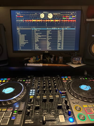 The image captures an indoor DJ setup from a high-angle perspective, showing a computer monitor, a DJ controller, and a studio monitor. The scene is illuminated by artificial light, suggesting an evening or nighttime setting.

The main subjects are the DJ equipment. On the left, a large monitor displays the Serato DJ Pro software interface, showing waveforms, track information, playlists, and a song titled "10% (feat. Kali Uchis) (Explicit)" by Kaytranada. Key elements like BPM (111.7 and 110.0), elapsed time (00:36.2, 02:54.8, 03:15.5), and track details are visible on the screen, which also bears the "ASUS" brand logo.

To the right, a Pioneer DDJ-FLX10 performance DJ controller dominates the frame, with numerous illuminated buttons, knobs, and faders. An Apple Watch rests on one of the jog wheels. The controller features sections labeled "PAD MODE," "HOT CUE," "CUE," "PLAY/PAUSE," "BEAT FX," "REVERB," "SAMPLER," "MASTER," and "TEMPO." A "KRK" brand studio monitor is partially visible in the upper background.

On the desk between the monitor and the controller, various small items are arranged, including a drumstick labeled "pro-mark," several small figurines or toys, and a decorative box. The overall impression is a dedicated home studio or DJ workspace.