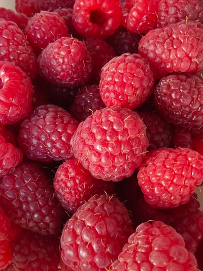 The image displays a close-up, overhead view of a large pile of ripe raspberries. The berries are primarily a vibrant red, with some appearing slightly darker or with hints of pink. They are clustered together, creating a textured surface. Each raspberry is composed of numerous drupelets, giving them their characteristic bumpy appearance. Fine, wispy hairs are visible on the surface of some of the berries. The lighting suggests an indoor setting, with highlights and shadows defining the shapes of the fruit. There are no people, animals, or recognizable structures visible. The exact location in Poltavka, Ukraine is not discernible from the image itself; it solely depicts the fruit.