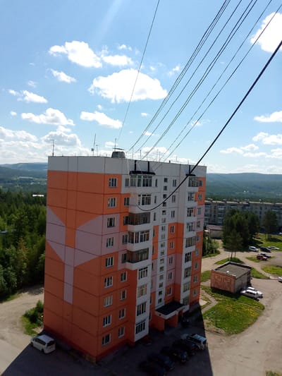 This is a high-angle, sunlit exterior shot of a multi-story apartment building in Neryungri, Russia, on a clear day with scattered clouds. The building, painted with an orange and light pink geometric pattern, dominates the foreground. Power lines stretch diagonally across the sky and over the building, obscuring parts of the blue sky.
Below and to the right of the main building, a smaller, brick structure with a grey roof is visible, adjacent to a patch of green grass dotted with yellow flowers. Several cars are parked in a paved area, including a white van, a white SUV, a red car, and several dark-colored sedans. A white minivan is parked on the street to the left of the main building.
The background features more apartment buildings of similar architecture, interspersed with evergreen trees and rolling hills. The overall scene depicts a residential area within the city. There are no people visible in the image.