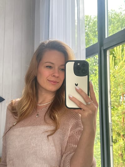 A woman is taking a selfie in a mirror. She is wearing a light pink, semi-sheer knitted sweater over a white top and a delicate necklace with a small shell pendant. She has long, blonde hair and is wearing a ring on her right hand, which is holding her phone. The phone case is off-white with a black border and features a black heart icon. The background appears to be an interior setting with a large window that looks out onto green foliage, suggesting it's daytime and the weather is pleasant. The reflection in the window indicates a bright, possibly sunny day. The woman has a gentle smile, conveying a relaxed and happy emotion. The setting seems to be a home, possibly in Znamenka, Russia.