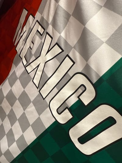 A close-up shot of a "Mexico" jersey. The fabric is divided into three sections, red, white and green, with white and gray checkered patterns on the white section. The word "Mexico" is printed diagonally across the fabric in large, bold letters with black outlines.