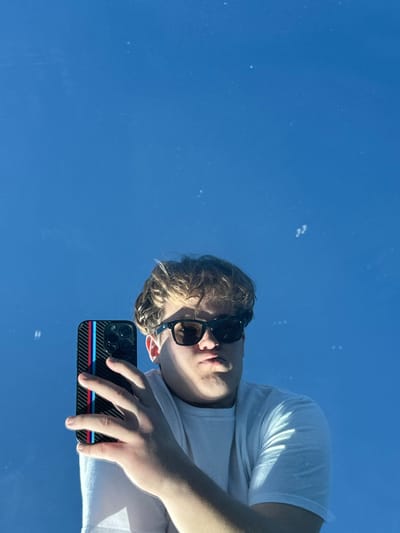 A man with sunglasses and a white t-shirt holds a phone with a carbon fiber case and blue, red, and white stripes. The photo is taken at an angle against a clear blue sky. The man's face and the phone are in focus, while the sky serves as a bright backdrop. The reflection on a surface may be responsible for this perspective.