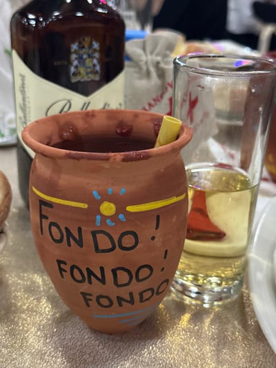 A brown, earthenware pot with "FONDO!" painted on its side in black lettering, accented with yellow and blue stripes and a sunburst design, sits next to a partially full glass and a bottle of liquor. The pot's interior appears to contain a dark, chunky substance. The scene is set on a textured, possibly metallic surface.