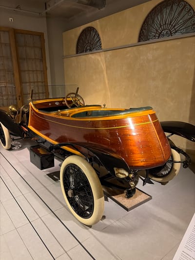 A vintage, boat-shaped car with a highly polished, reddish-brown wooden body is the central subject of the image, displayed indoors, likely in a museum in Wassenaar, Netherlands. The car features black fenders, a black chassis, and distinctive cream-colored, white-walled tires with black spoked wheels. Gold or brass accents are visible, including a steering wheel and a horn near the front wheel. The car's unique wooden construction, resembling a boat hull, is a prominent detail, with small fasteners or rivets visible across its surface and a yellow stripe running along its side.
The setting consists of light beige/tan walls with a textured finish and a light grey tiled floor with dark grout lines. Architectural details include two decorative wrought iron grilles with spiral motifs, one partially visible at the top left and another smaller one below. A large wooden window frame with multiple small panes is visible in the background. The car is presented on a low platform, with an information signboard standing next to it.
The visible text on the signboard, partially in Dutch and English, references "Panhard," "Nederlands" (Dutch), "houten plank" (wooden plank, referring to the body), and describes the vehicle as a "Boat-shaped body" and a "MASTERPIECE THAT set the trend." The text also mentions "In the 10" and "In de ja," indicating historical context. The environment suggests an exhibit, with artificial lighting illuminating the vehicle.
