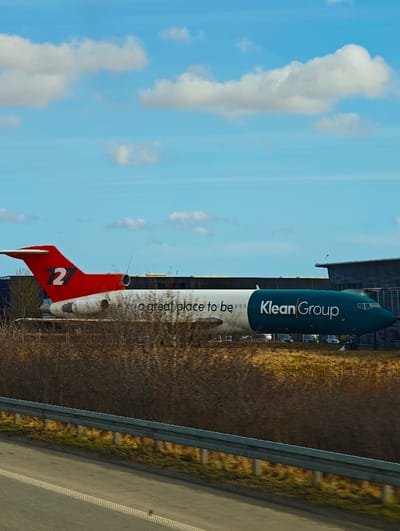 The image displays a static Boeing 727 aircraft, repurposed and located at the Klean Group headquarters in Skanderborg, Denmark. The aircraft's livery consists of a red tail section marked with a white "72" logo. The mid-fuselage is white, featuring black text that reads "a great place to be." The forward fuselage and nose cone are teal, bearing the white text "Klean Group." The plane is positioned on ground level, adjacent to a dark, modern corporate building that is partially visible behind it. In the immediate foreground, a multi-lane asphalt highway is visible, separated from the aircraft by a silver metal guardrail and a dense strip of brown, leafless deciduous vegetation. The sky above is clear blue with scattered white cumulus clouds. No individuals, specific actions, or interactions are present in the scene.