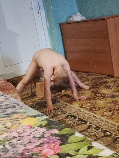 A young child is positioned on a carpet in what appears to be an indoor setting. The child is bent over backward in an arched position, using a small stool for support. The surrounding environment includes furniture, specifically a dresser, and an ornate carpet. The location is in Chernik, Bulgaria.