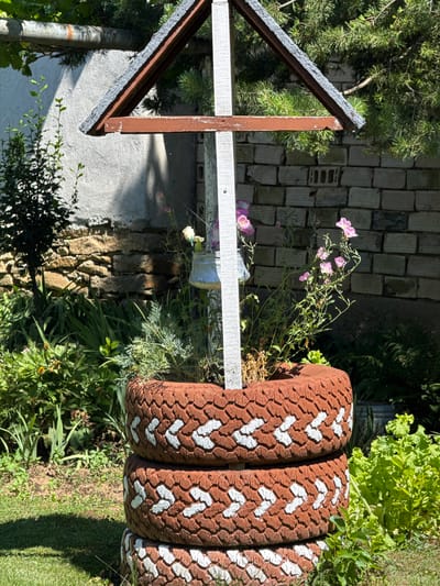 The image shows a decorative garden feature made from three stacked, painted tires. The tires are painted reddish-brown with white chevron patterns around their sides. In the center of the tires, a small wooden structure with a peaked roof has been built. Various plants, including green foliage and some pink flowers, are growing out of the top tire. A small metal watering can hangs from the wooden structure. In the background, a brick wall and a stone wall are visible, along with a large tree. The scene is set in an outdoor garden. The location is Konop, Bulgaria.