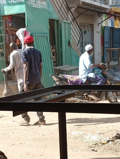 The image depicts an outdoor urban scene in Kofar Ruwa, Nigeria, under daylight conditions. In the foreground, a dark metal structural frame partially obstructs the view of the ground, which is unpaved, dusty, and shows tire tracks and scattered refuse.
Three men are visible. On the left, a man in a light-colored, long-sleeved garment holds a long-handled tool resembling a shovel, with its rounded blade resting on the ground, as he walks towards a green corrugated metal shop. Next to him, a man seen from the back wears a red baseball cap, a dark blue short-sleeved collared shirt with white trim, and dark trousers, walking towards the right side of the frame. In the center-right, a man rides a motorcycle from right to left. He wears a white skullcap, a light blue long-sleeved shirt, dark trousers, and a light green face mask. The motorcycle features a patterned seat covering.
In the background, two commercial establishments are present. The left building is a green corrugated metal shop with an open entrance, displaying goods inside. Signage above reads "NA JEGA" and includes the phone number "07046757310". To its right, another building, possibly made of concrete, features signage reading "MASIRA GLOBAL CONCEPT" and has an external metal staircase attached to its facade. Further right, a blue metal door is visible.