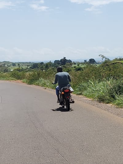 A man wearing a blue shirt and blue pants rides a black motorcycle down a paved road. The motorcycle has a white bag attached to the rear fender. The road is lined with tall green grass on both sides. In the background, there are rolling hills covered in trees and vegetation, with a hazy sky above. The scene appears to be in a rural or semi-rural area.