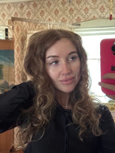 A selfie-style portrait features a young woman with long, curly light brown hair, appearing to hold a pink smartphone with an Apple logo above her head to capture the image. She has light-colored eyes and is looking directly at the camera with a neutral to slight smile. She wears a dark, possibly black, long-sleeved top and a silver ring on her left hand.
The background reveals an indoor setting, likely a room. Prominently featured is light-colored wallpaper with a recurring floral or geometric pattern in shades of beige and reddish-brown. A bright window is visible in the upper background, indicating daytime and providing natural light. Below the window, a portion of what appears to be a framed landscape painting or picture, featuring blue and green tones, can be seen. Another faint yellow, possibly floral, pattern is visible near the top right of the frame, behind the phone. The overall ambiance suggests a domestic interior in Ledukhi, Russia.