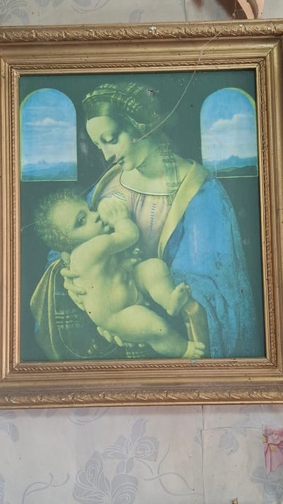 A framed print hangs on a wall, depicting a woman holding a baby. The artwork has an aged and somewhat faded appearance. The frame is ornate and gold-colored. The wallpaper has a floral pattern. The location is Шешурга, Russia.