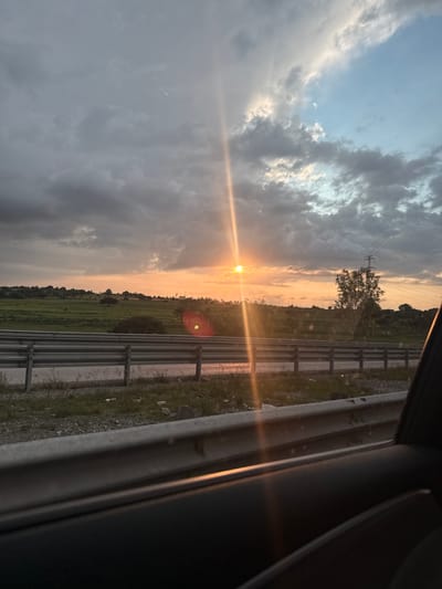 The image captures a sunset view from a vehicle on a highway in Coyotepec, Mexico. The sun is low on the horizon, casting a warm orange glow across the sky and illuminating a line of trees and power lines in the distance. Dark, ominous clouds fill the upper portion of the sky, contrasting with patches of lighter, clearer sky. A metallic guardrail, typical of highway infrastructure, runs horizontally across the foreground, partially obscuring a grassy embankment littered with debris. The interior of the vehicle is visible on the right side of the frame, with a dark dashboard and door panel. The overall scene depicts a dramatic and slightly moody sunset over a semi-rural landscape.