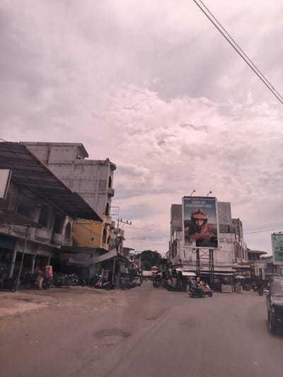 The image displays a daytime street scene in Meukokuthang, Indonesia, under an overcast sky with light, scattered clouds. The paved road, which appears somewhat worn, stretches into the distance, bordered on both sides by a mix of multi-story buildings and ground-level shops.

Several people are visible, mostly engaged with motorbikes; some are riding, while others are parked near the shops. Motorbikes are a prevalent mode of transport, with numerous scooters parked along the street. A car is partially visible on the far right foreground.

A prominent feature is a large billboard located on the right side of the street, showcasing an image of a person wearing a red motorcycle helmet. The billboard carries the Indonesian text: "USAHA TAK MENGKHIANATI HASIL" (Effort does not betray results) at the top. Below the main image, the text "ADA ADALAH Surya" is visible, followed by smaller print that likely constitutes a health warning, suggesting a cigarette advertisement for the "Surya" brand.

On the left side of the street, a tall, multi-story building with a raw concrete finish and arched openings is noticeable. Overhead utility wires crisscross the sky above the street. The scene portrays typical daily life and commercial activity in a moderately busy town environment, with people commuting and gathering near shops.