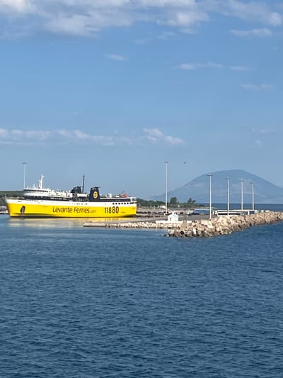 The image depicts a large yellow ferryboat, identified as "Levante Ferries," docked in the Municipal Unit of Kastro - Kyllini, Greece. The ferry has the number "11880" displayed prominently in yellow. The boat is situated in a harbor, with a breakwater made of large rocks extending into the blue water. In the background, there are several tall lampposts and a distant, hazy mountain. The sky is blue with scattered white clouds, indicating a clear day. The scene appears to be a port area, with no people clearly visible in the frame. The ferry is the primary subject, suggesting a transportation hub or a scenic view of maritime activity.