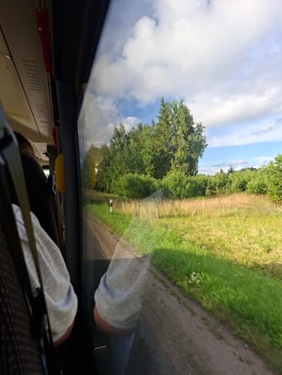 The image, taken from inside a vehicle, shows a reflection of the outside scenery in the window. The scene outside is a rural landscape with green trees and fields under a cloudy sky. A dirt path runs alongside the grassy area. The time of day appears to be daytime, with bright lighting and shadows suggesting it's not midday. The weather seems pleasant, with a mix of sun and clouds. Inside the vehicle, parts of a seat and the interior are visible, along with the reflection of a person's arm wearing a grey sweatshirt. The overall mood is calm and observational, as if someone is looking out the window during a journey. There is no visible text in the image.