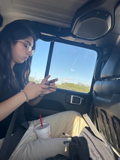 Captured from within a vehicle, the photograph reveals a young woman with dark, wavy hair and glasses, focused on her cell phone. She is seated adjacent to another person whose tan pants and drink with a red straw are visible. The scene unfolds against a backdrop of a bright, cloudless sky and distant greenery, presumably viewed through the car window. The interior of the vehicle features a black speaker on the upper left and various car components. The location is Comfort, United States of America.
