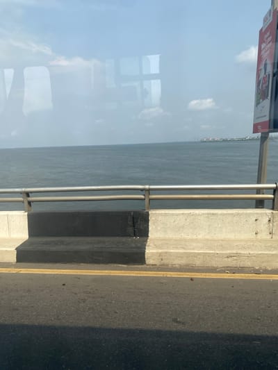 The image presents a roadside perspective from an elevated structure, likely a bridge, overlooking a large body of water near Ebute-Metta, Nigeria. The immediate foreground features a section of asphalt road with a solid yellow lane marking. A dark, elongated shadow crosses the lower left portion of the road surface. Adjacent to the roadway is a concrete barrier, with its left segment painted matte black and its right segment remaining unpainted light gray concrete. Above this barrier, a metal guardrail system, composed of two parallel horizontal bars supported by vertical posts, runs along the edge of the structure.
Beyond the guardrail and barrier, a vast expanse of dark blue-gray water, appearing choppy, extends to the horizon. In the far distance across the water, a landmass is visible, featuring a low-rise skyline with several buildings and distinct tall, slender structures, possibly industrial chimneys or communication towers. The sky above is a light blue with scattered white clouds, appearing somewhat hazy. Reflections of window panes, forming a grid pattern, and other interior vehicle elements are superimposed across the upper portion of the image, indicating the photograph was taken through glass. A partial billboard on a vertical pole is visible on the far right, displaying a figure in a red shirt, text including "Global No. 1 brand," and the logo "TCL." No persons or specific dynamic actions are discernible within the frame.