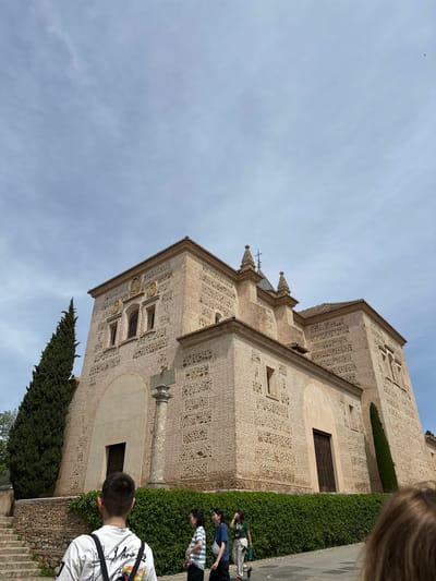 The image presents a tilted view of a historic building, likely a church or monastery, under a bright, partly cloudy sky. The structure, characteristic of Southern Spanish architecture, features facades built with light-colored bricks arranged in intricate, decorative patterns, possibly reflecting Mudejar design influences. The building exhibits multiple levels with arched and rectangular windows, along with ornamental crests or medallions embedded in the brickwork. Terracotta tiles cover sections of the roof, and decorative spires or finials adorn its silhouette.
Several people, appearing to be visitors or tourists, are visible in the foreground and bottom right. One person, seen from behind, wears a white long-sleeved shirt with partial text "MONA" and other letters (possibly "MONARCHY ART") printed on the back. Behind them, a small group includes individuals in dark blue and green shirts. The top right corner shows the hair of another person, positioned very close to the camera.
A tall, dark green coniferous tree, likely a cypress, stands prominently to the left of the building. A well-maintained green hedge runs along the base of the structure, separating it from a paved path where the people are walking. A stone cross on a pedestal is situated next to the building, indicating its religious significance. Steps are visible in the bottom right corner. The sky is a clear blue with scattered white clouds, signifying daytime. A small dark bird can be seen flying high in the sky. The architectural style strongly suggests the location is in Granada, Spain, depicting a historical landmark.