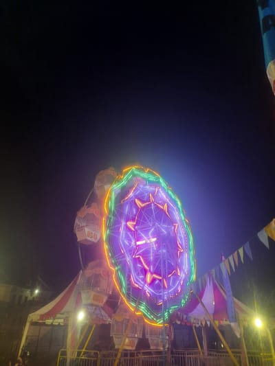 This forensic photograph depicts an outdoor carnival scene at night in Sukasari, Indonesia. A large Ferris wheel, illuminated with bright neon purple, green, and orange lights, dominates the center-right of the frame. Several gondolas or carriages are visible on the Ferris wheel, with "04," "05," and "06" distinctly marked on some. Beneath the Ferris wheel, carnival tents with red and white stripes, as well as pink and purple sections, are erected. Metal railings forming a barrier are present in the foreground. Overhead, multicolored triangular bunting is strung across the scene. The immediate area around the tents and Ferris wheel is illuminated by additional yellowish artificial lights. The background consists of a dark night sky, with indistinct outlines of distant buildings faintly visible on the far left. No identifiable individuals are present within the captured scene.