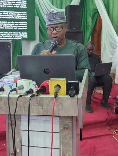 The image captures a man, presumably giving a speech, standing behind a podium with multiple microphones bearing logos of various news outlets, including "Nigerian Television," "Trust," and "Arise News." He is wearing a traditional cap and glasses, holding a microphone in his right hand, and facing a laptop with an HP logo. Another man sits in the background with a laptop on his lap. The podium is cluttered with wires and microphone attachments. A green and white draped cloth hangs in the background. The scene suggests a press conference or public event in Ungwan Liman, Nigeria.