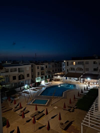 The image depicts a nighttime view of a resort or hotel complex in Paralimni, Cyprus. On the left, the sky transitions from dark blue to a lighter hue near the horizon. Adjacent to the sky is a multi-story building with several windows, some of which are illuminated. To the right of the building is a pool area with a large swimming pool and a smaller adjacent pool. Several red beach umbrellas are scattered around the pool, casting shadows on the paved area. The pavers are composed of different shades. Lush greenery is present near the pool area. A white railing is visible at the top right of the image. The overall impression is of a quiet evening at a resort.