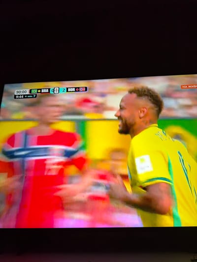 The image is a television screen displaying a football match with a score overlay. On the left side of the overlay, it shows "90:00", the Brazilian flag, "BRA", a score of "0", a Norwegian flag, and "NOR" with a score of "2". Below this, it reads "9:44 + min. 7". On the right side of the screen, a man in a yellow jersey with green trim is visible in profile, smiling. He appears to be a football player. Another player in a red jersey with blue and white stripes is blurred on the left. The background is out of focus, showing stadium lights and blurred spectators. A red banner with white Cyrillic text, "НА ЖИВО", is visible in the top right corner of the screen, indicating "LIVE" broadcast. This scene is a television broadcast and not a real-world location in Ognianovo, Bulgaria.