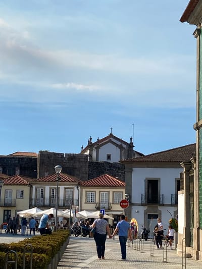 The image depicts a bustling street scene in Vila Nova de Cerveira, Portugal, on a clear day with scattered white clouds. In the background, the prominent white facade of a church, featuring a central cross and two finials, is visible atop dark stone fortification walls, characteristic of Cerveira Castle. Below these fortifications, a row of traditional Portuguese buildings with light-colored facades, dark-framed windows, and red terracotta roofs line the street.
The midground shows a cobblestone public area with multiple individuals. To the left, several people are seated at outdoor cafe tables under white parasols. One man in a light blue shirt is bent forward, looking down at an object near a planter. To the right, a building with a corner visible features a distinctive green-tiled exterior. A red circular "STOP" sign is mounted on a pole near this building. Parked motorcycles are visible in the vicinity of the cafe area. Further right, colorful, abstract sculptures, one resembling a large green insect, are arranged along the path.
In the foreground, a couple walks away from the viewer, holding hands, along the cobblestone path. The woman wears a dark shirt and jeans, while the man wears a blue shirt and jeans. The path is flanked by rectangular planters containing green shrubbery and metal barrier posts. Several other pedestrians, including children, are dispersed throughout the scene.