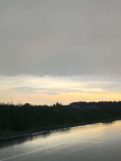 This media file captures a serene rural landscape in Paderina, Russia. The scene is dominated by a vast, overcast sky, with a band of soft, pale yellow and orange light near the horizon, suggesting either sunrise or sunset. Below the sky, a dense line of dark green foliage runs horizontally across the frame, punctuated by the silhouettes of houses and trees. A tall, slender antenna is visible in the distance to the left, amidst the greenery. The foreground features a wet asphalt road, reflecting the muted light and the overcast sky, with a clear white line marking the edge of a lane. The wetness of the road indicates recent rainfall, contributing to the overall peaceful and slightly melancholic atmosphere of the scene. No people or specific objects are distinctly visible beyond the natural elements and structures. The time of day is suggested by the soft glow on the horizon, likely early morning or late evening. The overall impression is one of quiet natural beauty in a rural setting.