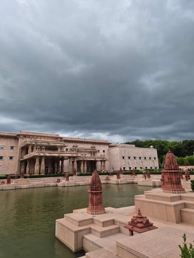 The image captures the exterior of a grand stone building, likely a temple or cultural center, in Robbinsville Township, United States of America. The architecture is ornate, with multiple levels, intricate carvings on pillars and facades, and a covered walkway supported by columns. In the foreground, a body of greenish water, possibly a reflecting pool or pond, is visible, with tiered stone platforms and decorative red spires emerging from the water and the surrounding stone structures. The sky above is heavily overcast with dark, dramatic clouds, suggesting an impending storm or recent rainfall. A few people are sparsely scattered in the background near the building entrance, and trees line the far edge of the property. The overall scene conveys a sense of solemnity and grandeur, amplified by the moody sky.