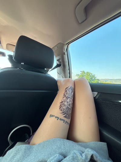 The image depicts the interior of a vehicle with a person's legs visible in the foreground. The person has a tattoo on their left thigh that appears to be of a skull and flowers. There is also text tattooed below the image on the thigh. The person is wearing grey sweatpants. The interior of the car includes a dark-colored headrest and seat. Through the car window, a bright blue sky and green trees are visible, suggesting an outdoor environment. The scene appears to be a snapshot taken during a journey, possibly on a road trip, and is not specifically located in Valkovo, Bulgaria.