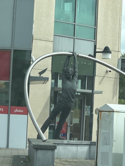 A dark bronze statue of a man hanging from a metal archway is displayed in front of a modern building with large windows. The man is depicted with his arms extended and gripping the arch, with his body in a bent position. The building appears to be a commercial establishment, with signage visible on the windows, including "PS" and "Sodas." The statue is situated on a concrete platform, which is placed on a paved area. In the background, a lamppost with a dark cap is visible, and a security camera is mounted on the building. The overall scene suggests an urban public art installation. This statue is known as the "George Bernard Shaw Sculpture" and is located in Carlow town, County Carlow, Ireland.