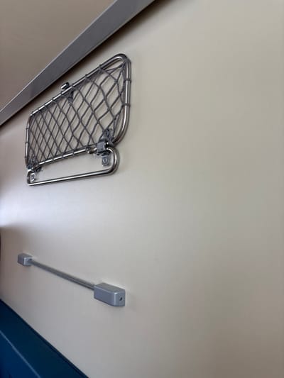 The image depicts an interior view of a compartment, likely within a modern transport vehicle such as a train. The primary surface is a smooth, light beige or cream-colored wall that occupies most of the frame. Along the upper left edge of the wall, a polished metallic trim piece, appearing to be stainless steel or aluminum, runs horizontally.
Mounted on the wall in the upper-left quadrant is a polished stainless steel frame containing a gray, coarse-mesh net, serving as a luggage or storage net. Directly below this net, a separate, hinged stainless steel bar is attached horizontally.
Below the luggage net and slightly to its right, another horizontal utility fixture is present. This consists of two rectangular, silver-colored end mounts attached to the wall, between which a thin, coiled or flexible silver-colored bar extends.
At the bottom-left edge of the frame, a small section of a dark blue surface is visible, consistent with the top edge of an upholstered seat or bench. No individuals, actions, or specific interactions are present in the scene. The lighting is even, originating from the left. The image provides no visual information to determine a specific geographical location within Ivanovka, Russia or any other city.