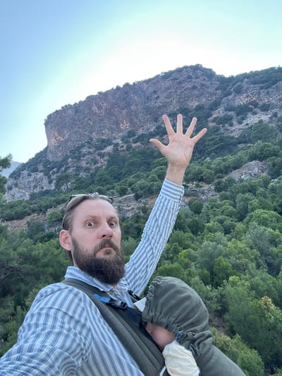 A man with a beard and glasses on his head is taking a selfie while carrying a baby in a carrier. The man is wearing a blue and white striped shirt. He has his left hand raised, showing five fingers. Behind him, there is a large, rocky mountain with many carved-out tombs or caves visible on its face, surrounded by lush green trees. The sky is a clear light blue. This scene is located in the area of Seydikemer, Turkey, specifically at the ancient Lycian rock tombs of Tlos.
