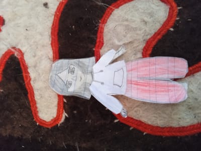 A drawing of a paper doll of a young person is placed on a textured surface with red yarn interwoven into it. The doll has short, gray hair and is wearing a white jacket with a pocket and pink checkered pants. The doll's arms are outstretched, and its legs are bent. The background appears to be a rug or blanket with areas of white, black, and brown fibers, along with the red yarn. The image is a close-up shot, and the lighting is neutral, suggesting an indoor setting. There is no visible text in the image. The overall impression is of a handmade craft or toy.