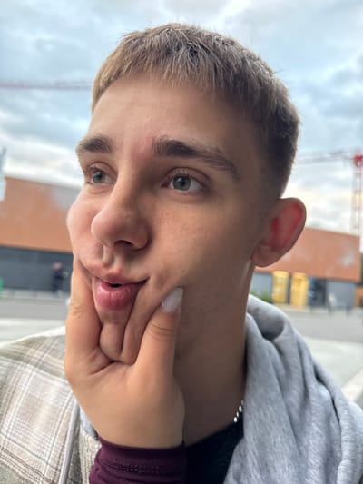 A close-up, self-taken photograph depicts a young adult male with short, blond hair, looking to his left with his cheeks puffed out and his lips pursed. His left hand is pressed against his right cheek, creating a distorted facial expression. He is wearing a plaid shirt over a grey hooded sweatshirt. In the background, a blurred urban environment is visible, featuring buildings and construction cranes. The image does not provide any specific information to determine its location within the city of Lörrach, Germany.