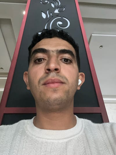 A medium shot portrait shows a man wearing a white knitted sweater. He has dark, curly hair and brown eyes. He is wearing white wireless earbuds in his right ear. The man is facing forward and looking directly at the camera. Behind him is a dark triangular structure with a red border, featuring a white decorative pattern. The background appears to be an indoor setting with neutral-colored walls and ceiling fixtures, suggesting a modern interior. There is no indication of the specific location within Beni Mellal, Morocco.
