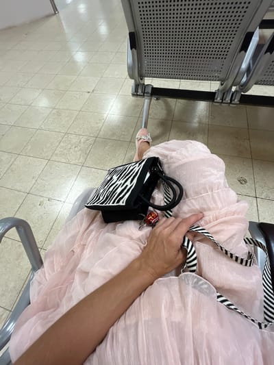 An overhead shot captures a person's hand resting on a light pink, flowing dress. The dress drapes over a person's lap as they sit in a gray metal waiting area chair. A black and white zebra-print handbag is placed on the dress. The bag has two black handles and a black and white striped strap. A small red and gold keychain hangs from the bag. The person's right arm is visible, extending from the lower left of the frame. Their feet, clad in light pink sandals, are visible in the upper left portion of the image, resting on a beige tiled floor. In the background, more gray waiting chairs with perforated seats are visible. The scene appears to be an indoor waiting area, possibly at an airport in Katunayake, Sri Lanka, given the architectural style of the chairs and the tiled floor commonly found in such public spaces.